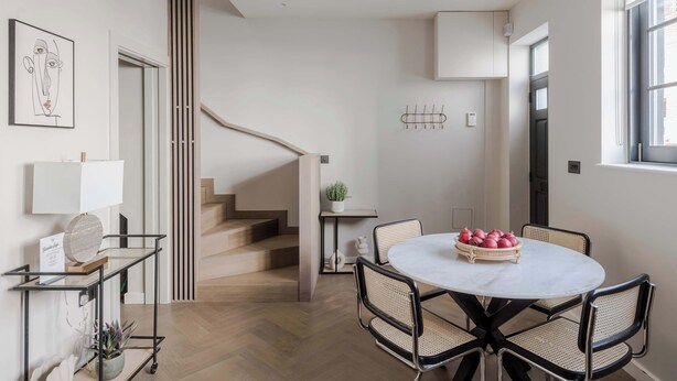 A modern interior with a staircase, a round dining table with chairs, a small side table with a plant, and a wall-mounted light fixture. - Marylebone Mews House · Regent's Park · Sleeps 6 (London)