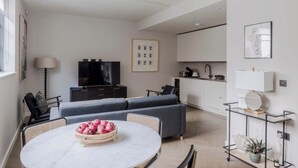 A modern living room with a sofa, a dining table with a fruit bowl, a TV, and a kitchen area with cabinets. - Marylebone Mews House · Regent's Park · Sleeps 6 (London)
