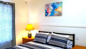 Ein Schlafzimmer mit einem Bett, gestreiften Kissen, einem Nachttisch mit Lampe, einem Bild an der Wand und einem Fenster mit Vorhang.