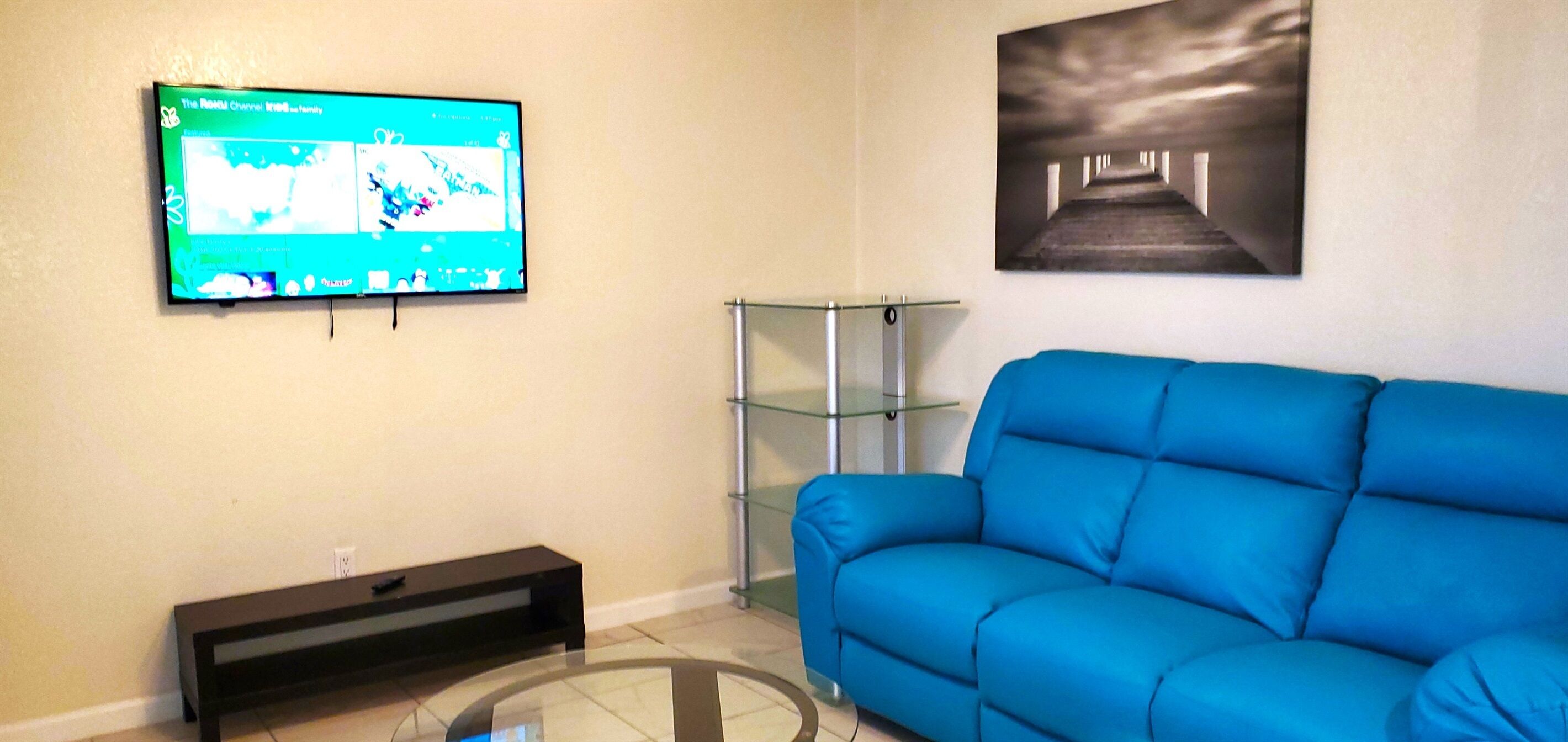 Un salon avec un canapé bleu, une table basse en verre, un téléviseur fixé au mur et un tableau au-dessus du canapé.