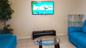 Ein Wohnzimmer mit flachbildfernseher an der Wand, einem Glastisch und einem blauen Sofa sowie einer schwarzen Bank.