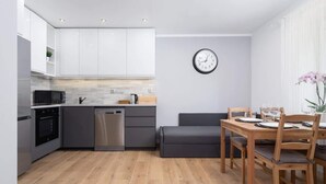 A modern kitchen with white cabinets, a microwave, oven, and dishwasher. There's a dining area with a wooden table and chairs, and a gray sofa.