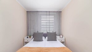 A bedroom with a bed, two bedside tables, a nightstand, a window with blinds, and a wall-mounted plant.