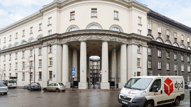 Ein großes, klassizistisches Gebäude mit einem zentralen Eingang, der von Säulen gesäumt wird, einem Parkplatzschild und einem weißen Lieferwagen mit rotem Logo.