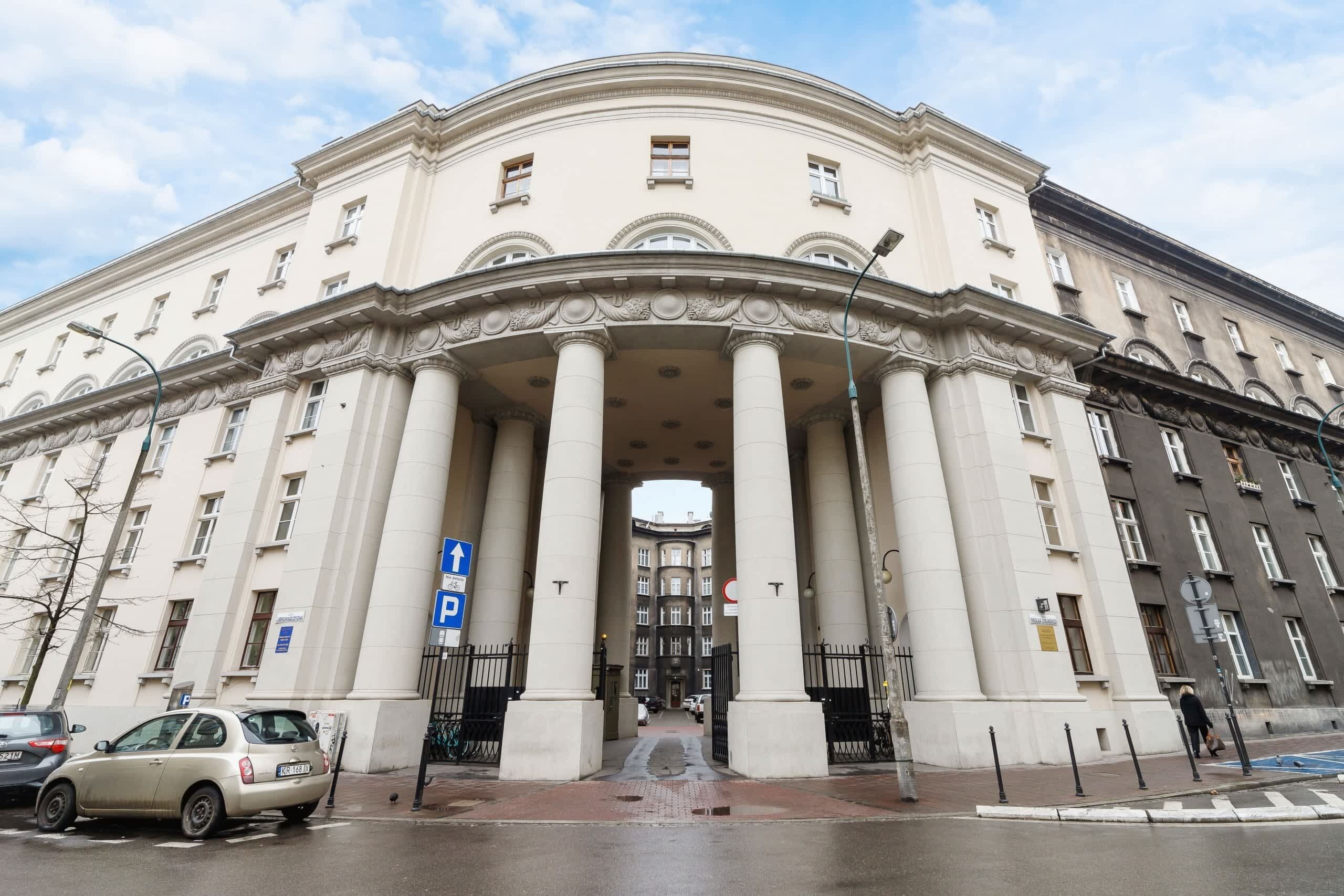 A large, classical building with a central entrance flanked by columns, a blue parking sign, and a pedestrian crossing.