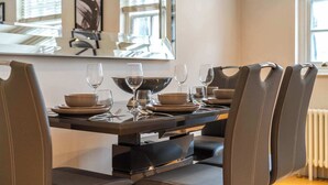 Un coin repas avec une table dressée pour six personnes, des chaises, un grand miroir et une fenêtre avec des stores.