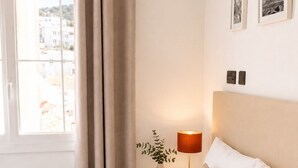 A neatly made bed with a beige bedspread and pillows, a rolled white towel, a bedside lamp, and a window with curtains. - Grand Central House - Penthouse Double Room (Gibraltar)