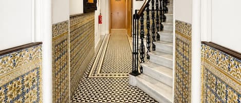 A hallway with ornate white arches, a patterned tiled floor, and a staircase with black railings.