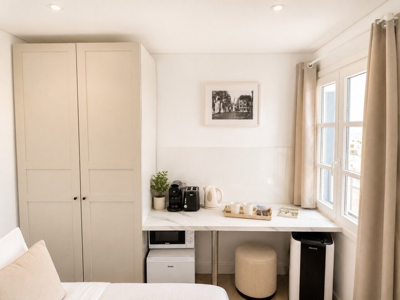 A bedroom with a bed, a desk with a coffee maker, a trash bin, and a window with curtains.