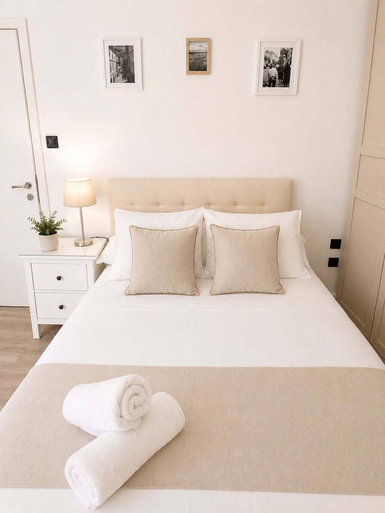 A neatly made bed with a beige headboard, two pillows, a rolled white towel, and a bedside table with a lamp and a potted plant.