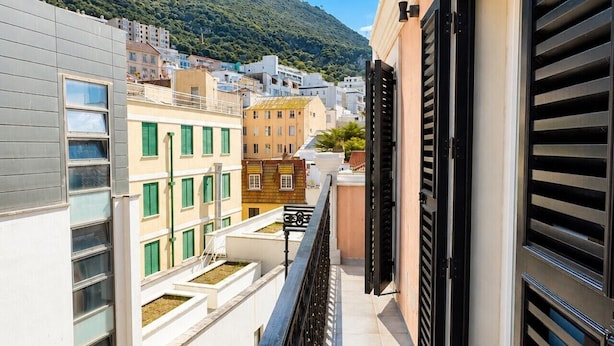 Un balcón estrecho con barandilla metálica que da a una vista urbana con edificios y una colina al fondo.