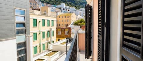 Un balcón estrecho con barandilla metálica que da a una vista urbana con edificios y una colina al fondo.