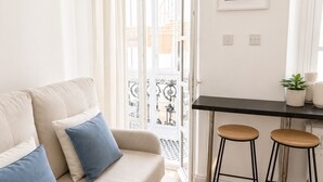 A living room with a sofa, two blue cushions, a dining table with two stools, and a framed picture on the wall. - Grand Central House - Classic One Bedroom Balcony (Gibraltar)