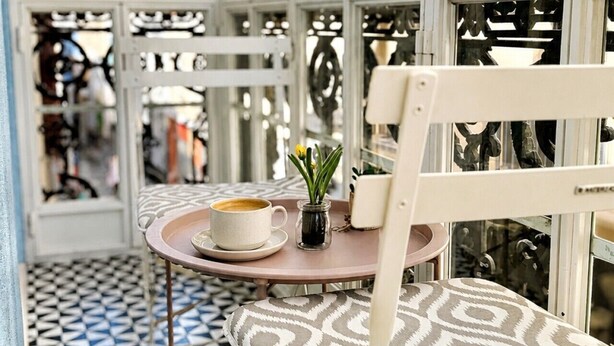 A sunlit outdoor seating area with a patterned floor, a table with a cup and a small plant, and a white chair. - Grand Central House - Classic One Bedroom Balcony (Gibraltar)