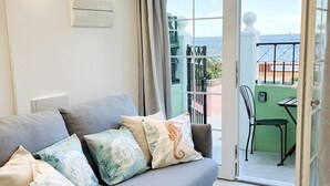 Un saló modern amb un sofà gris, coixins estampats, una porta de vidre que dona a una zona exterior i una taula petita amb una cadira.