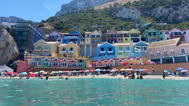 Un complex d'edificis costers amb diverses estructures de colors, una platja al primer terme i un turó rocós al fons.