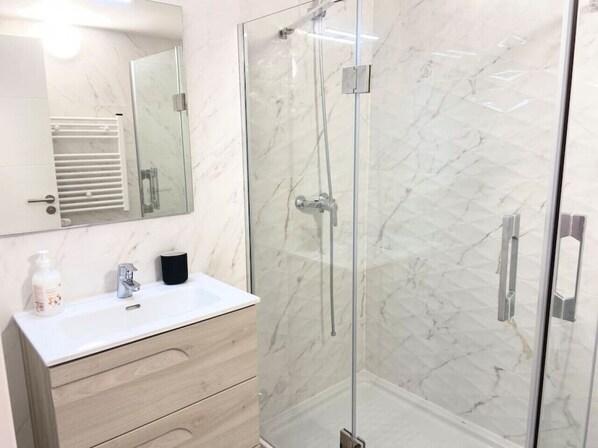 Een moderne badkamer met een douchecabine van marmer, een witte wastafel met spiegel erboven en een houten opbergmeubel met lades.
