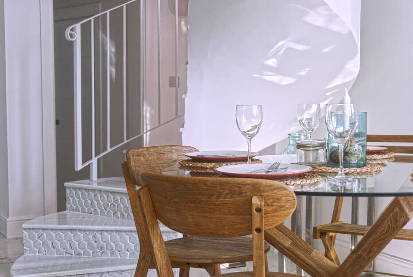 Un comedor con una mesa de vidrio, sillas de madera y una escalera con escalones blancos.