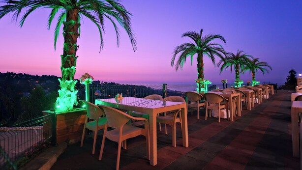 Un espace de restauration en plein air avec des tables et des chaises, des palmiers et une vue sur la ville au crépuscule.