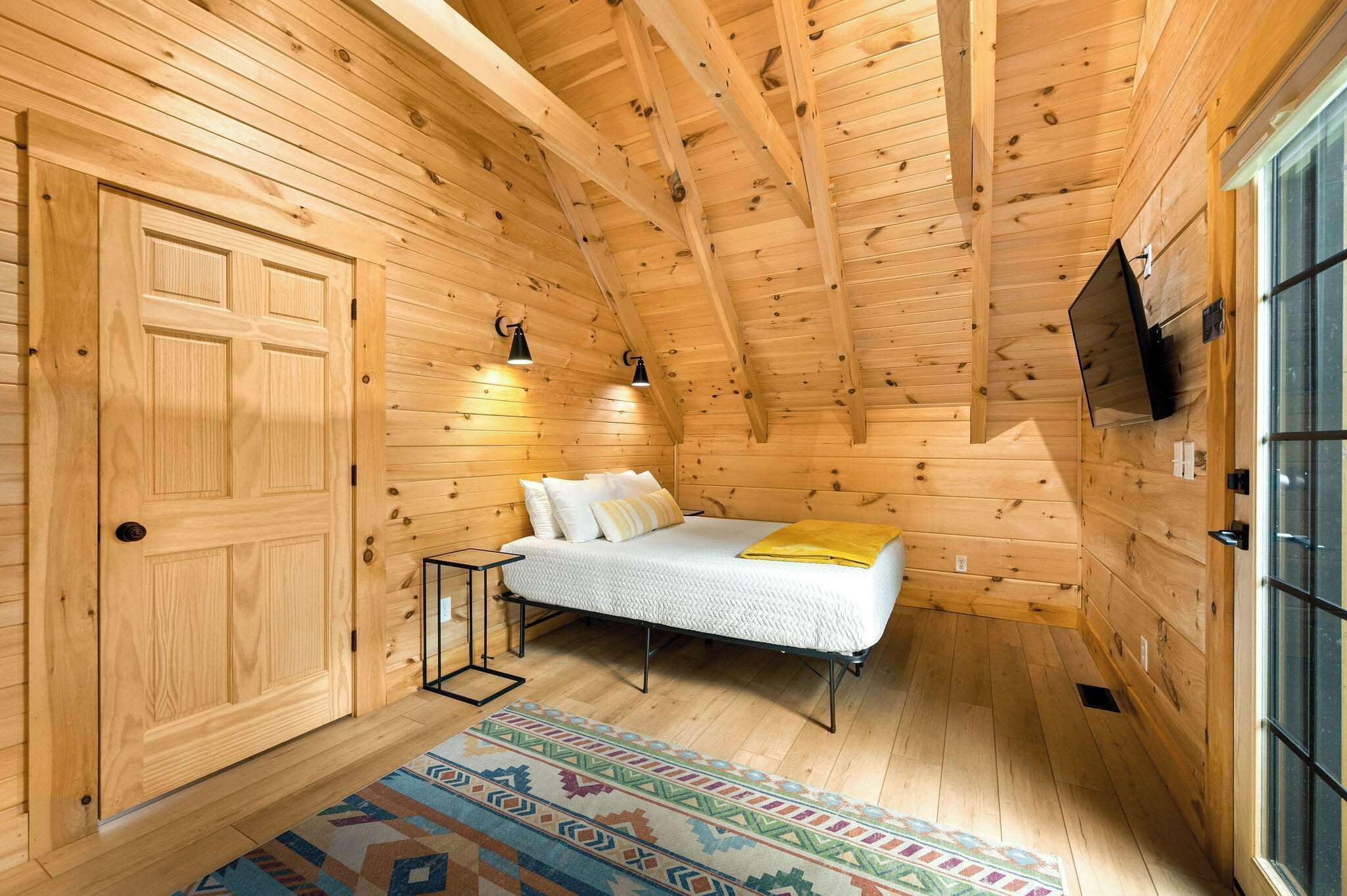 A wooden cabin bedroom with a bed, a nightstand, a TV mounted on the wall, and a large window.