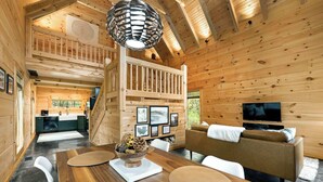 Ein Holzblockhaus-Interieur mit Essbereich, einem Wohnzimmer mit Sofa und Fernseher sowie einer Küche im Hintergrund.