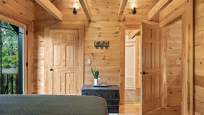 Un dormitorio de cabaña de madera con una cama, una maleta, una planta en maceta y una puerta que da a otra habitación.