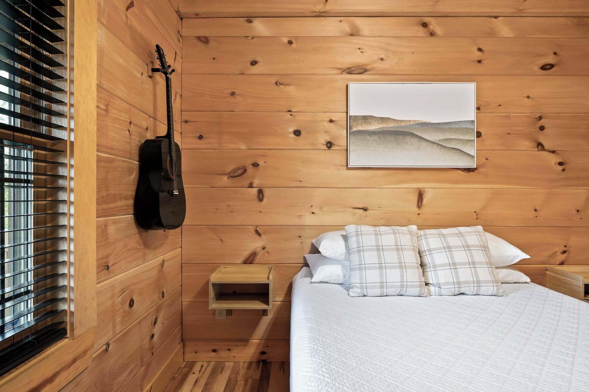 A wooden-paneled room with a bed, a guitar, a small wooden nightstand, and a painting of a landscape.
