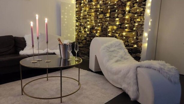 Un salón moderno con una pared de piedra, una mesa redonda con velas y un sofá blanco cubierto de piel sintética.