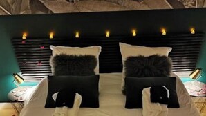 Una cama con un arreglo de pétalos de rosa en forma de corazón y velas, un cabecero oscuro y una pared con un estampado floral grande.