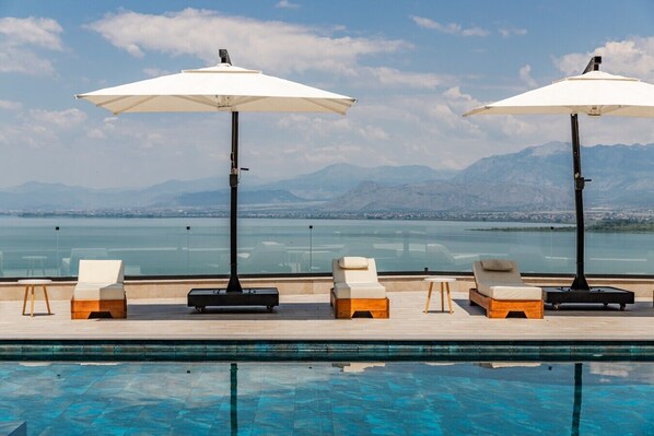 A poolside area with lounge chairs and umbrellas, overlooking a body of water and mountains.