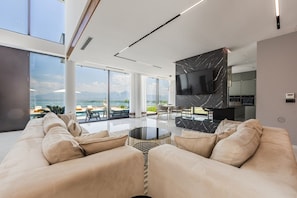 A modern living room with a large TV, comfortable seating, and a view of the sea.
