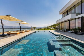 A modern pool area with lounge chairs and umbrellas, a clear view of the mountains, and a contemporary building with large windows.