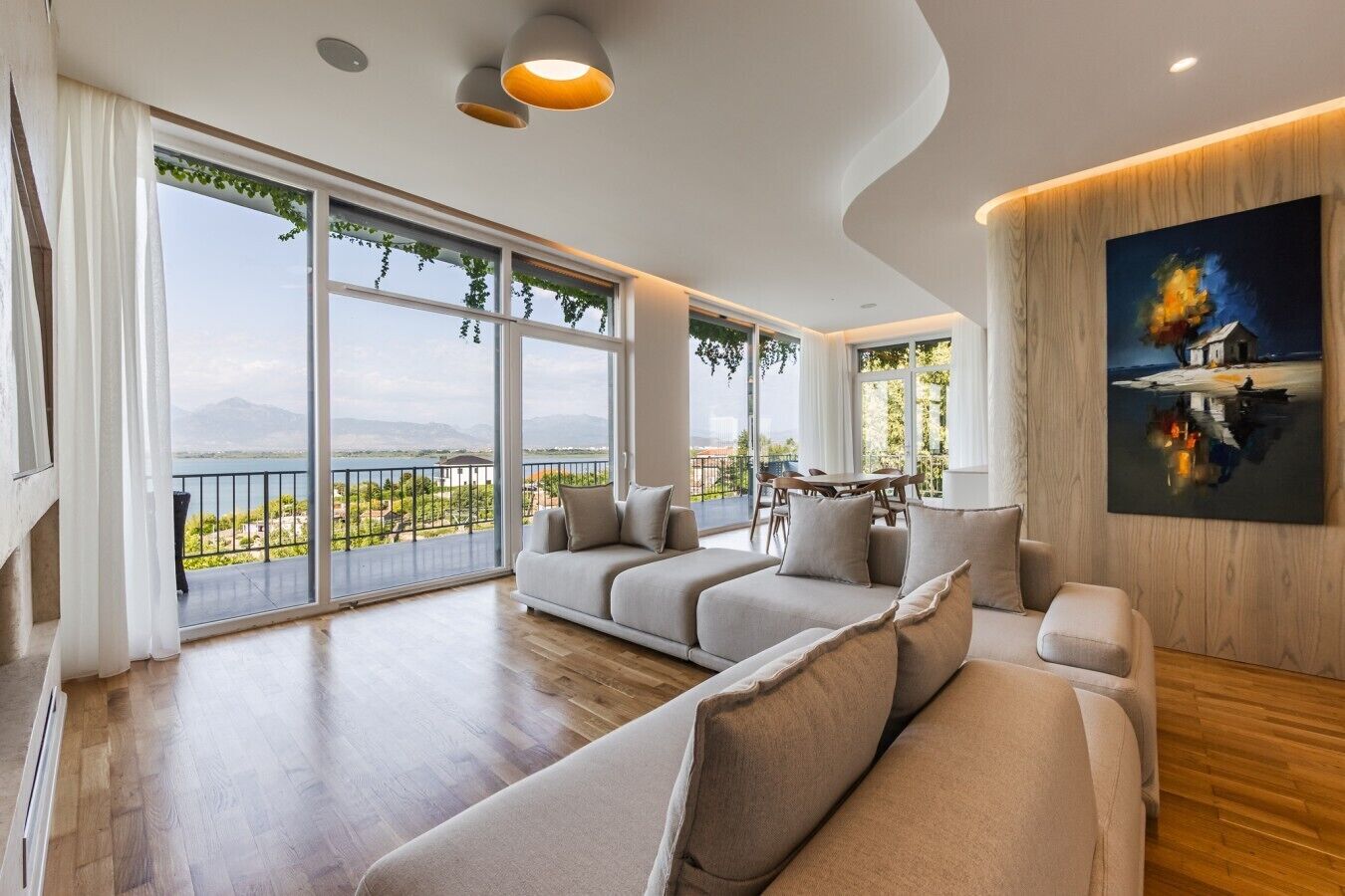 Ein geräumiges Wohnzimmer mit einer großen Couch, einem Esstisch und Blick auf ein Gewässer durch große Fenster.