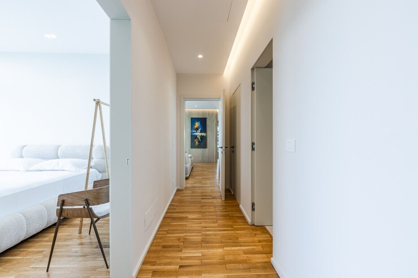 A modern hotel corridor with a wooden floor, white walls, and a bed with white bedding.