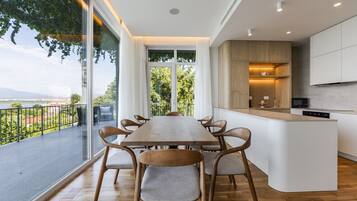 Een moderne keuken met een centraal eiland, een eethoek met een houten tafel en stoelen, en uitzicht op een wateroppervlak door grote ramen.