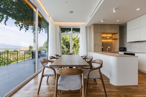 Une cuisine moderne dotée d’un îlot central, d’un coin repas avec une table et des chaises en bois, et offrant une vue sur un plan d’eau grâce à de grandes fenêtres.