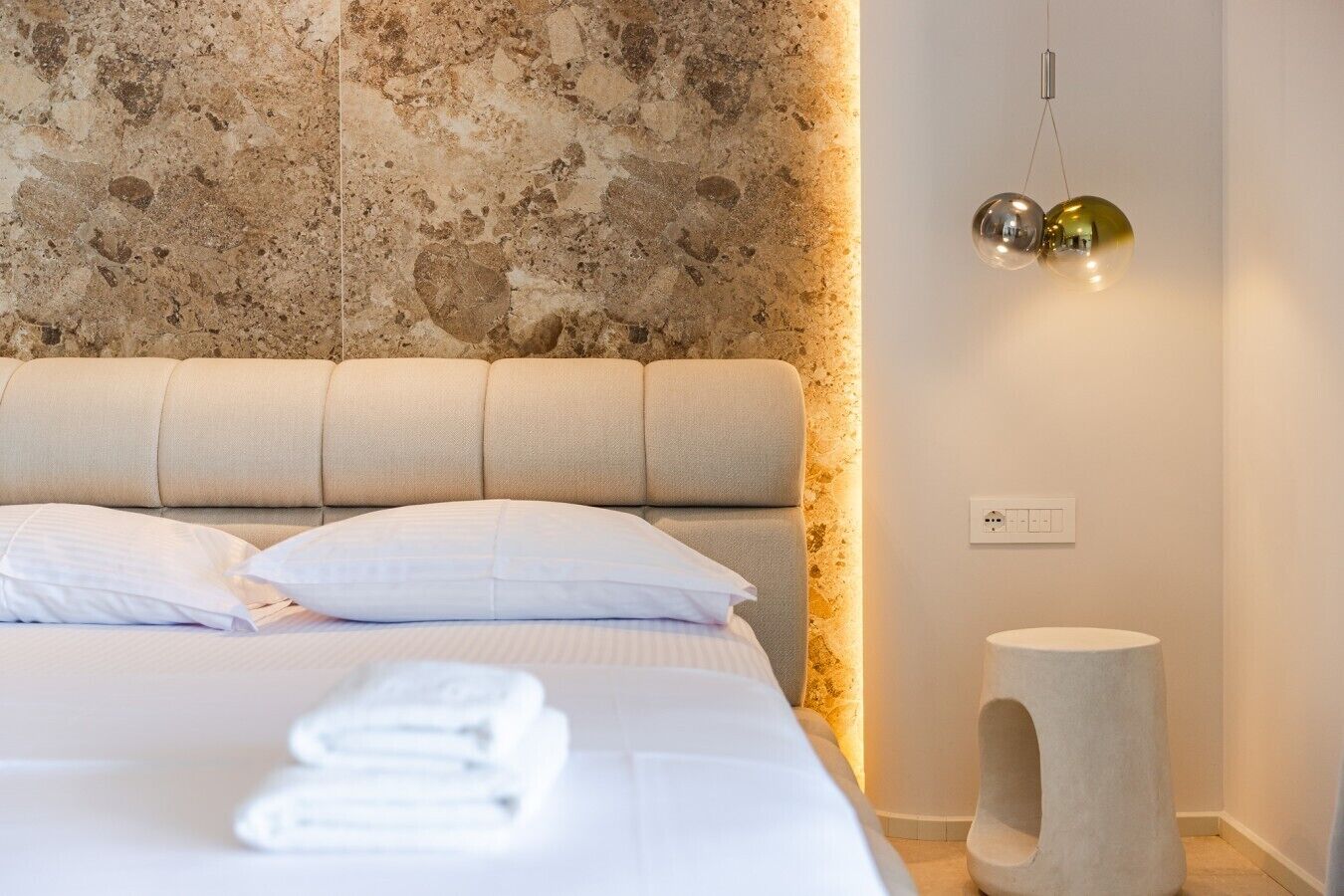 A neatly made bed with pillows and folded towels, a textured wall behind, a small round stool, and a pendant light.