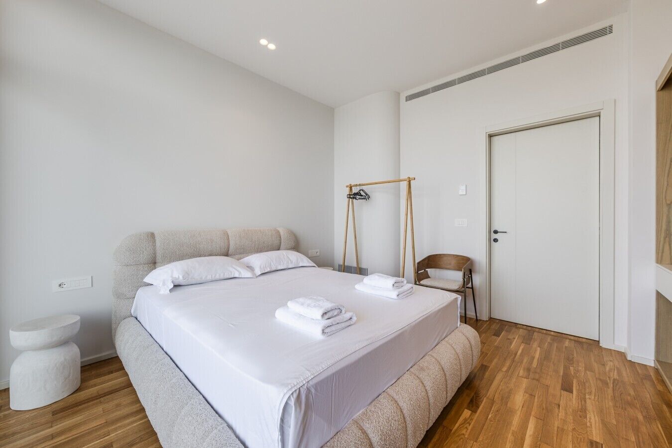 A modern bedroom with a large bed, a wooden floor, a white door, and a small chair.