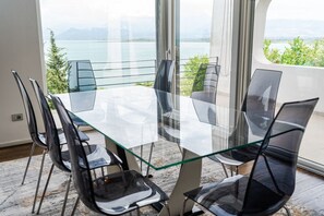 A modern dining area with a glass table and chairs, large windows offering a view of a lake and mountains.