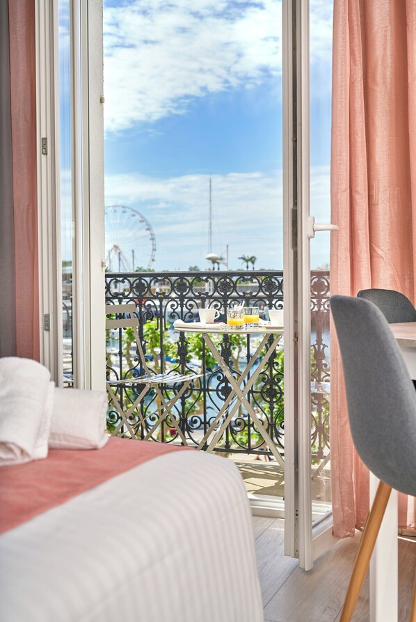 A hotel room with a bed, a table with a chair, and a view of a Ferris wheel through a balcony with a railing.