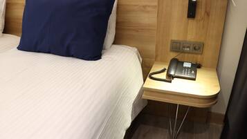 Una habitación de hotel con cama, almohada azul, mesita de noche con teléfono y cabecero de madera.
