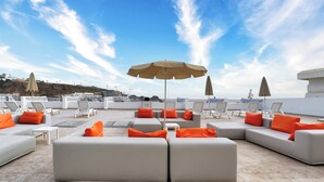 A rooftop lounge area with white sofas and orange cushions, outdoor umbrellas, and a view of the sea and buildings in the background. - Vistaflor Puerto Plata (Mogan)