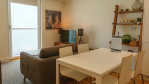 A modern living room with a sofa, a dining table, and a TV. There is a window with blinds, a painting on the wall, and a shelf with decorative items. - Still Life Canary Wharf Executive  (London)