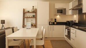 A modern kitchen with white cabinets, a built-in microwave, and a coffee maker. There is a wooden shelf with decorative items and a framed menu on the wall. - Still Life Canary Wharf Executive  (London)