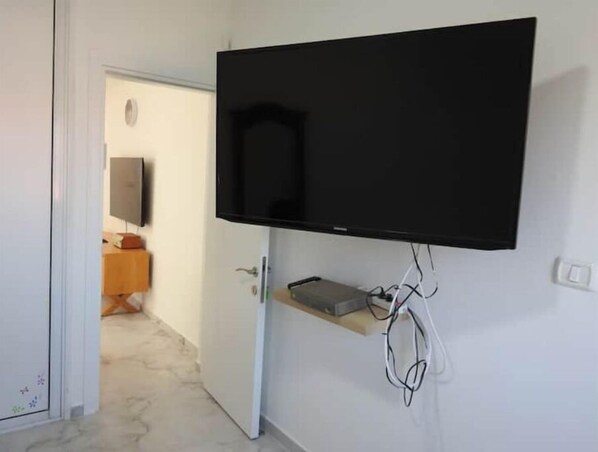 A room with a mounted flat-screen TV on the wall, a door leading to another room, and a small shelf with electronic devices.