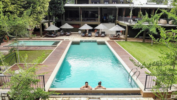 A rectangular swimming pool with two people at the far end, surrounded by wooden deck and stone walls.
