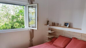 Un dormitorio con cama, ventana, mesita de noche y estante con objetos decorativos.