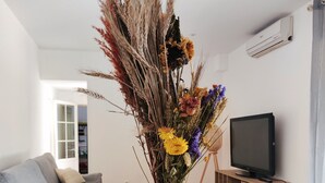 Una sala de estar con un jarrón de cristal con flores secas sobre una mesa redonda, un sofá gris y un televisor sobre un mueble de madera.