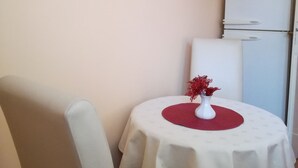 Een kleine kamer met een ronde tafel, een witte stoel, een witte koelkast, en een rode vaas met bloemen.