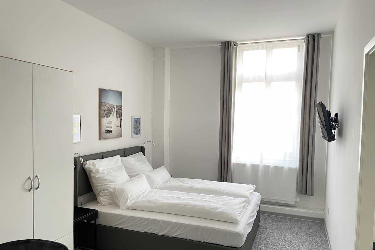 Quarto de hotel bem arrumado com cama, guarda-roupa, escrivaninha e janela com cortinas.